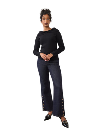 A model stands in a studio wearing a black long-sleeve top and dark blue wide-leg jeans with gold-tone button details near the hems, paired with black pointed shoes.