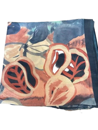 A folded patterned scarf or shawl laid on a carpeted floor, featuring an abstract botanical design with coral-red leaf shapes, cream and blue-gray areas, and a dark navy border. A person's feet in black sandals are visible at the bottom edge.