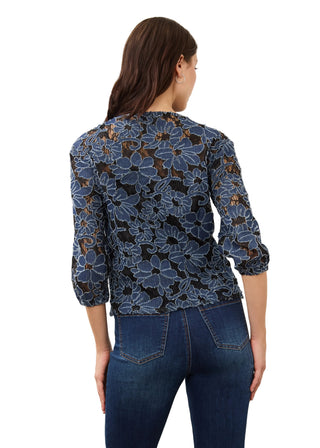 A woman viewed from the back wearing a dark blue floral lace top with sheer sleeves and dark blue jeans, against a plain light background.