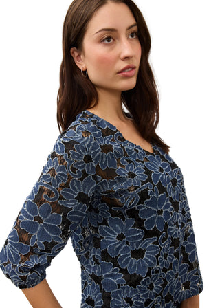 A woman wearing a blue floral lace top with three-quarter sleeves, shown from the chest up against a plain light background.