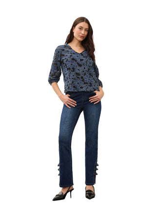 A woman modeling a dark blue patterned long-sleeve top and dark blue straight-leg jeans with decorative embellishments at the cuffs, paired with black pointed-toe heels, against a plain studio background.