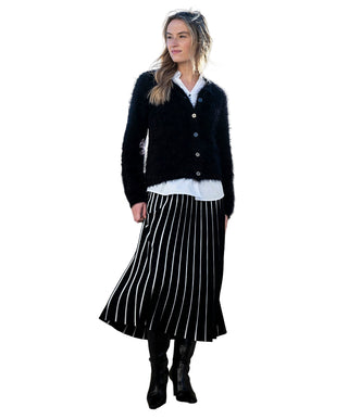 Woman wearing a black cardigan, white shirt, and black and white striped skirt against a white wall.
