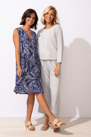 Two women standing side by side, one in a blue dress and the other in a white outfit, against a plain background.