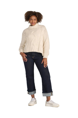 A woman modeling a cream cable-knit turtleneck sweater, dark blue cropped jeans with rolled cuffs, and white sneakers against a light gray studio background.
