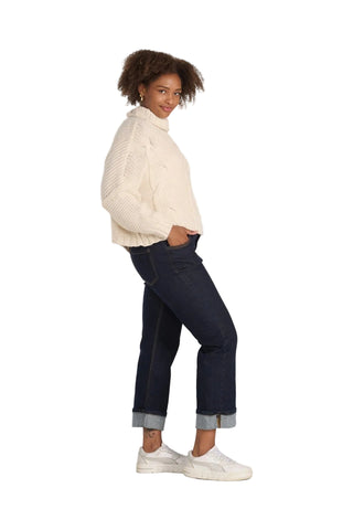 A woman modeling a cream cable-knit turtleneck sweater, dark blue cuffed jeans, and white sneakers against a light gray studio background.