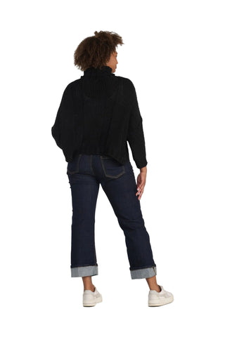 A woman is shown from behind wearing a black ribbed turtleneck sweater, dark blue cropped jeans with rolled cuffs, and white sneakers, set against a solid light gray background.