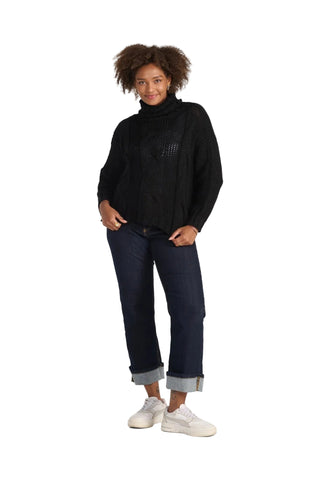 A woman modeling a black cable-knit mock neck sweater, dark blue jeans with rolled cuffs, and white sneakers against a light gray studio background.