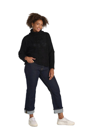 A woman modeling a black knit turtleneck sweater, dark blue jeans with rolled cuffs, and white sneakers against a light gray studio background.