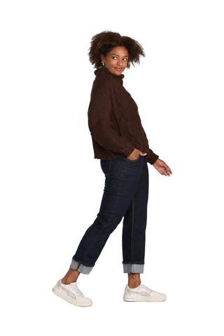 A woman modeling a dark brown knit turtleneck sweater, dark blue cuffed jeans, and white sneakers against a light gray studio background.