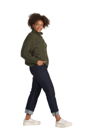 A woman modeling a dark olive green knit turtleneck sweater, dark blue jeans, and white sneakers against a solid light gray background.