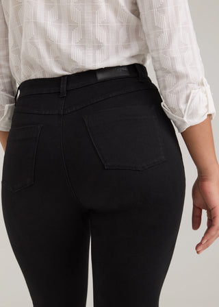 Close-up back view of a person wearing black high-waisted jeans with a matching black belt, paired with a white patterned blouse, against a light gray studio background.