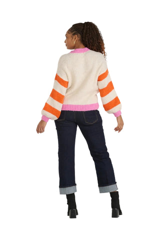 A woman shown from the back wearing a cream knit sweater with bold orange and pink horizontal stripes, paired with dark blue jeans and black ankle boots, against a light gray studio background.