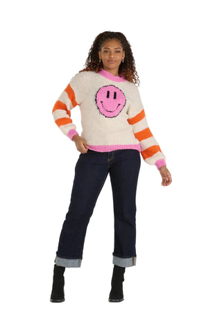 A woman modeling a cream-colored fuzzy knit sweater with orange and pink horizontal stripes, featuring a large pink smiley face on the front. She is wearing dark blue jeans with rolled cuffs and black ankle boots, standing against a light gray studio background.