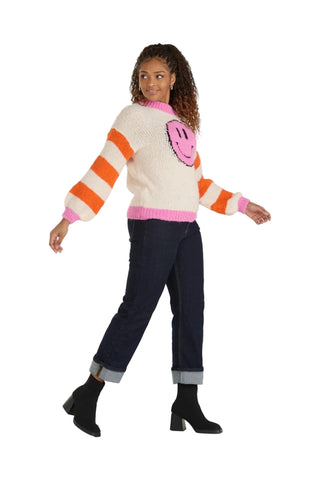 A woman modeling a cream knit sweater with orange and pink stripes, a pink smiley face motif on the chest, and a pink crew neck and hem, paired with dark blue jeans and black ankle boots against a light gray studio background.