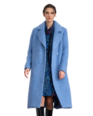 Woman wearing a long blue coat over a patterned dress in a room setting.