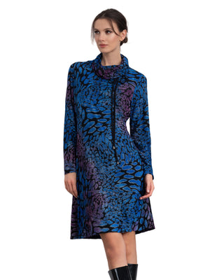 Woman wearing a blue and purple patterned dress against a white wall.