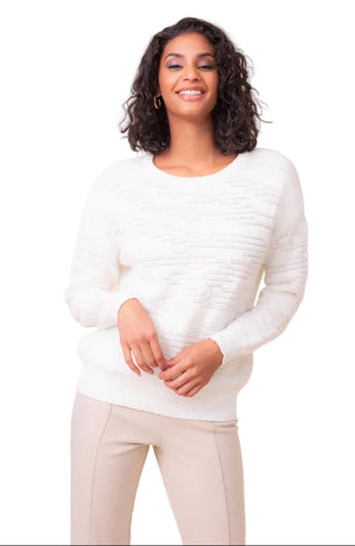 A woman with curly dark hair wearing a white textured knit sweater and beige pants, standing against a solid light beige studio background.