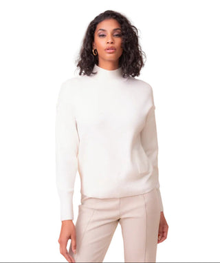 A woman with curly dark hair wearing a cream-colored mock neck knit sweater and light beige trousers, standing against a solid light beige background.