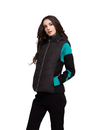 A woman modeling a black sleeveless puffer vest with teal and black color-blocked sleeves, paired with black pants, against a solid beige background.