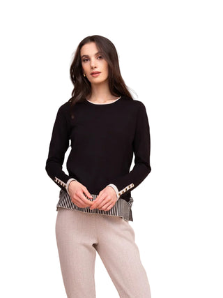 A woman modeling a black long-sleeve knit top with decorative white and black striped cuffs, paired with light gray pants, against a solid beige background.