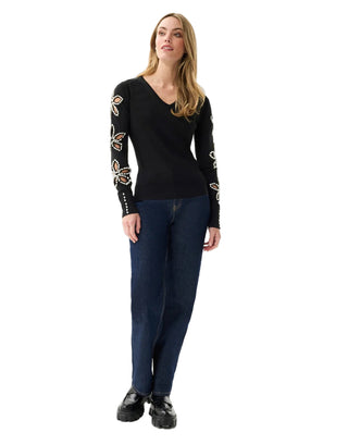 Woman wearing a black sweater with floral patterns on sleeves and dark jeans against a white background