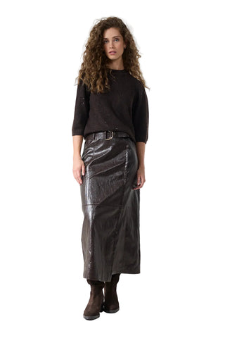 A model with curly brown hair stands against a white studio background wearing a dark brown knit sweater, a dark brown faux-leather midi skirt with a belt, and dark brown ankle boots.