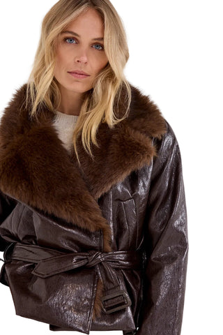 A woman wearing a dark brown faux-leather belted jacket with a large brown fur collar, photographed against a light gray studio background.