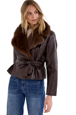 A woman wearing a dark brown faux-leather wrap jacket with a large brown fur collar and a matching tie belt, paired with blue jeans, against a light gray studio background.