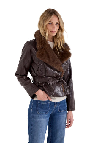 A woman modeling a dark brown faux-leather wrap jacket with a thick brown fur collar, belted at the waist, paired with blue jeans against a light gray studio background.
