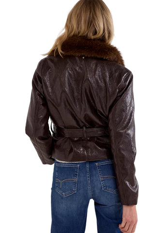 A woman viewed from behind wearing a dark brown glossy leather jacket with a brown fur collar and a matching belt, paired with blue jeans, against a light gray studio background.