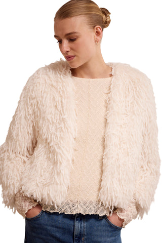 Woman wearing a cream-colored fluffy jacket over a lace top with blue jeans on a plain background