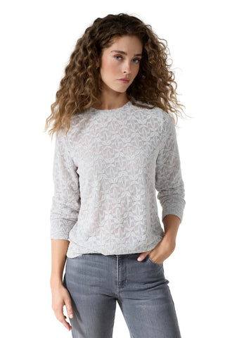 A woman with long, curly brown hair wearing a light gray textured knit sweater and gray jeans, standing against a plain white studio background.