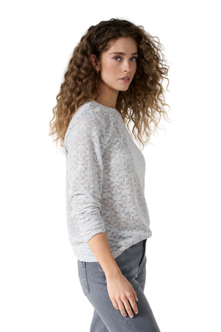 A woman with long, curly brown hair is shown in profile wearing a light gray and white patterned knit sweater and gray jeans against a plain white studio background.