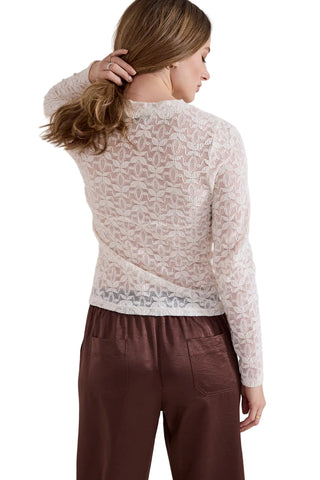 A woman shown from the back wearing a sheer cream lace long-sleeve top and brown pants against a light gray studio background.
