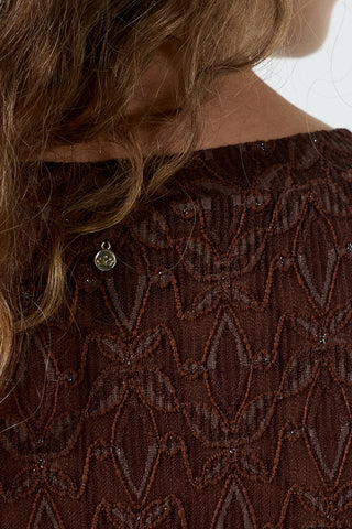 Close-up of a person wearing a dark brown lace top with a geometric pattern and a small round metallic charm at the neckline.