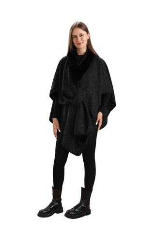 Woman wearing a black poncho over a black outfit against a white paneled wall.