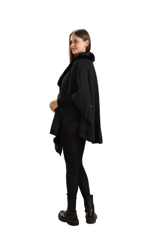 Woman wearing a black poncho and leggings standing on a wooden floor with a white paneled wall background