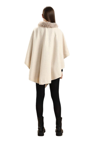 Woman wearing a cream-colored cape with fur trim against a white paneled wall.