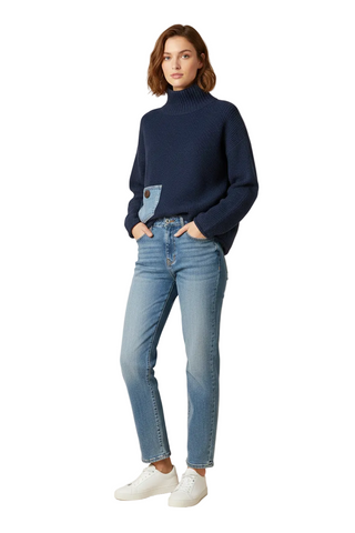 A woman stands against a plain beige studio background wearing a navy ribbed turtleneck sweater with a light blue patch pocket, light-wash blue jeans, and white sneakers.