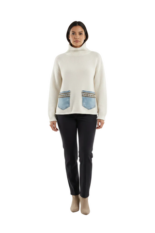 A model stands against a plain white studio background wearing a cream-colored knit turtleneck sweater with two light blue denim patch pockets featuring embroidered details, paired with black pants and beige ankle boots.