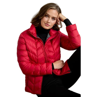 A woman wearing a bright red quilted puffer jacket over a black turtleneck and black pants, posing in a studio setting with a beige wall background.