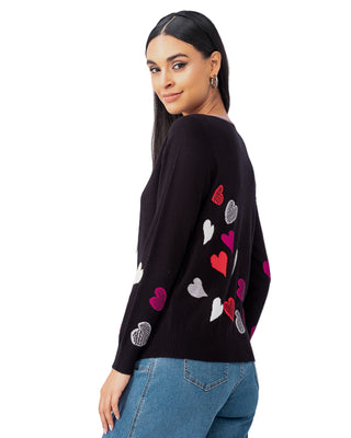 Woman wearing a black sweater with colorful heart patterns against a white curtain background