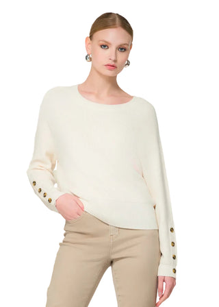 Woman wearing a white sweater with gold buttons and beige pants, standing next to flowers on a white background