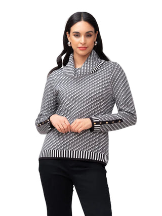 A woman modeling a black and white geometric patterned mock neck sweater with black trim and gold button accents on the cuffs, paired with black pants, against a light gray studio background.