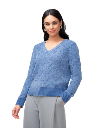A woman modeling a blue textured knit V-neck sweater with ribbed cuffs and hem, paired with light gray trousers, against a plain light gray studio background.
