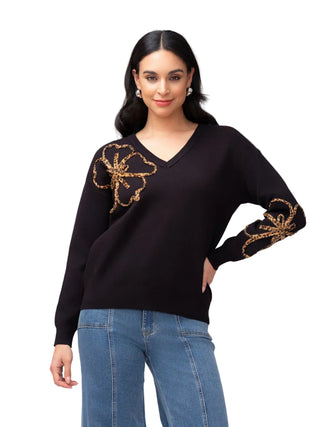 A woman modeling a black V-neck knit sweater with ornate gold embroidered floral motifs on the chest and sleeve, paired with blue jeans against a light gray studio background.