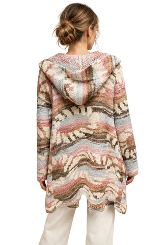 A woman viewed from behind wearing a loose, long-sleeve hooded knit cardigan with a multicolor Southwestern-style wavy pattern in pink, beige, brown, and light blue, paired with white pants against a light gray studio background.