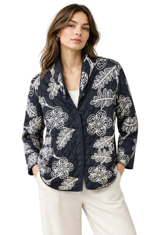 A quilted women's jacket with a black base and white floral and leaf pattern, hanging on a hanger in a retail store.