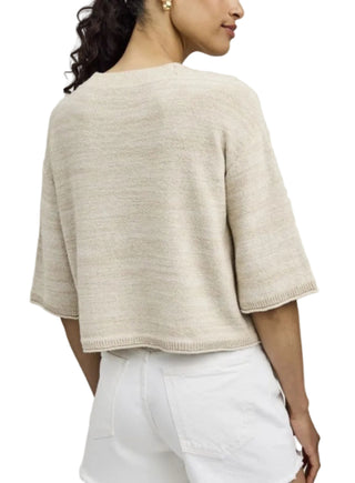 Woman wearing a beige knit top and white pants on a plain background