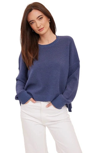 A woman wearing a ribbed knit crewneck sweater in a dusty periwinkle blue color, paired with white pants, against a plain light background.